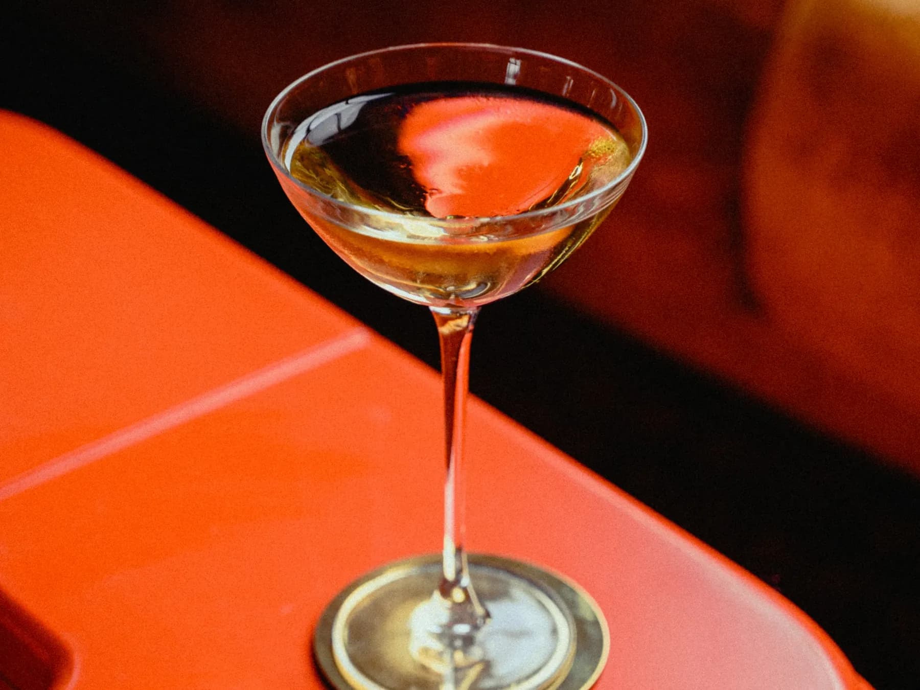 Golden Martini limpido e dorato in una coppetta a stelo lungo, su un sottobicchiere dorato e un piano arancione