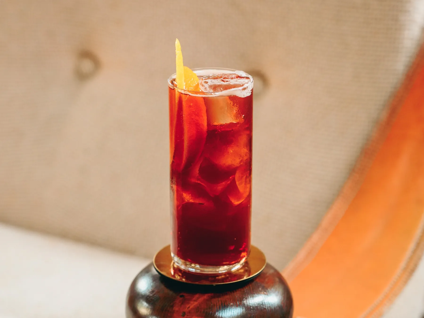 Golden Negroni rosso rubino in un bicchiere alto con ghiaccio, una fetta e una scorza d'arancia, su un sottobicchiere dorato