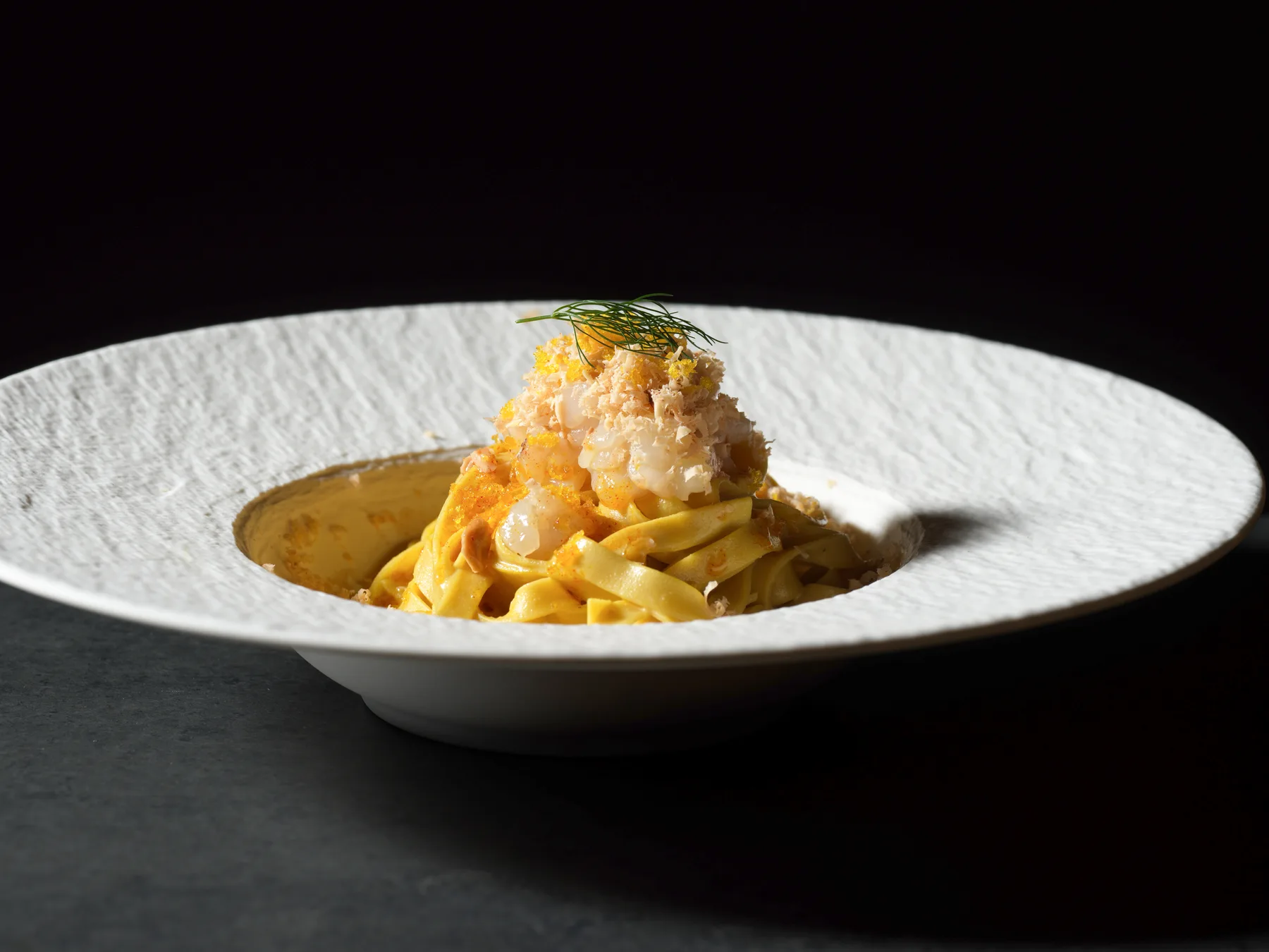 Taglioli allo zafferano con tartare di capasanta, mandorle tostate, bottarga e Lingotto grattugiati sopra, in un piatto bianco su fondo scuro