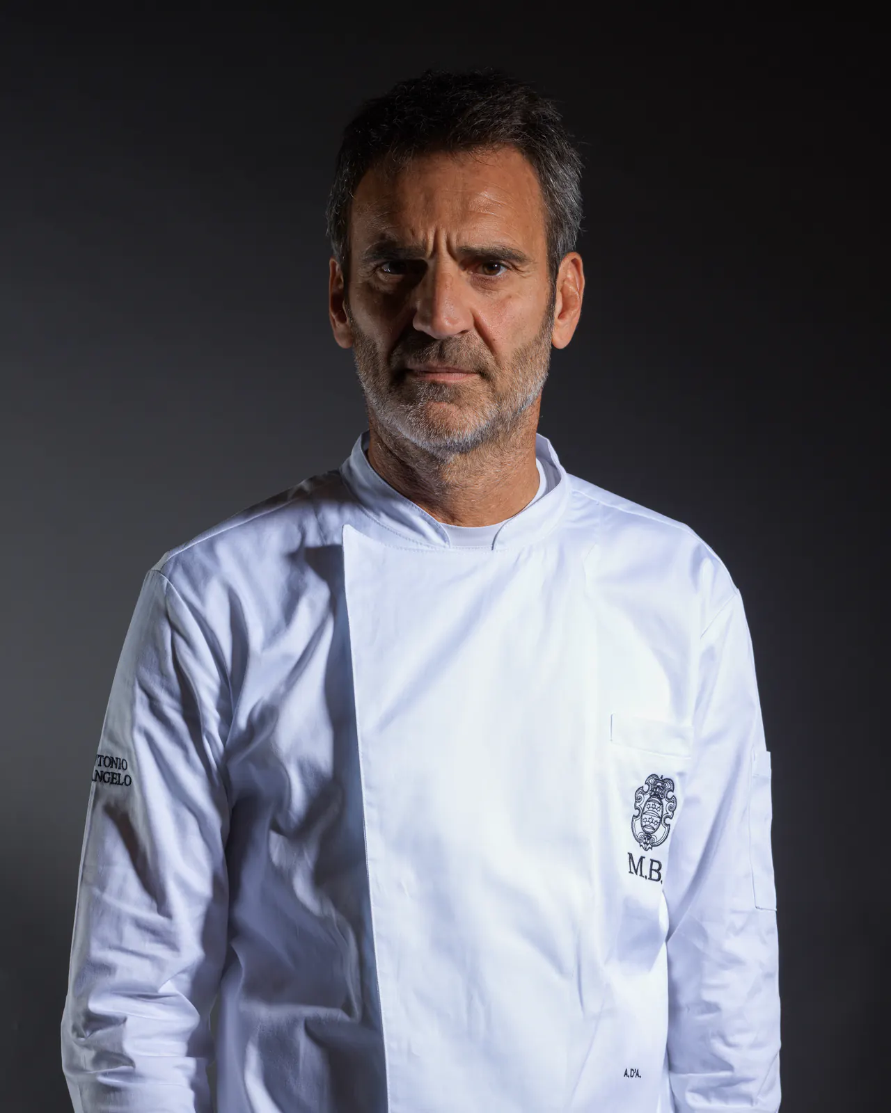 Antonio D'Angelo in giacca da cuoco bianca, con lo stemma M.B. ricamato sul petto, su fondo scuro